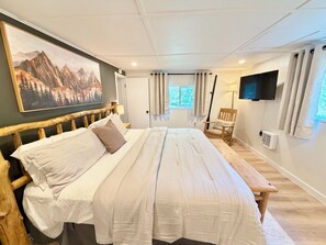 A bedroom with a large bed, a wooden headboard, a mounted TV, a window with curtains, a chair with a 'WELCOME' pillow, and a painting of mountains.