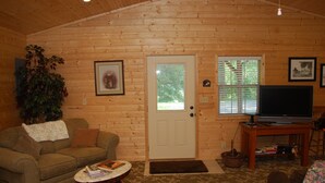 A cozy log cabin interior with a wooden door, a television on a wooden stand, a sofa, a small table, and a potted plant.