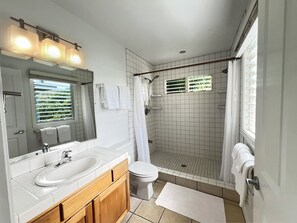 A bathroom with a white sink, mirror, and toilet, a shower with a glass door, and a window with blinds.