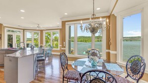Una cocina amplia con una isla grande, un comedor con mesa de vidrio y grandes ventanales con vistas a un lago.