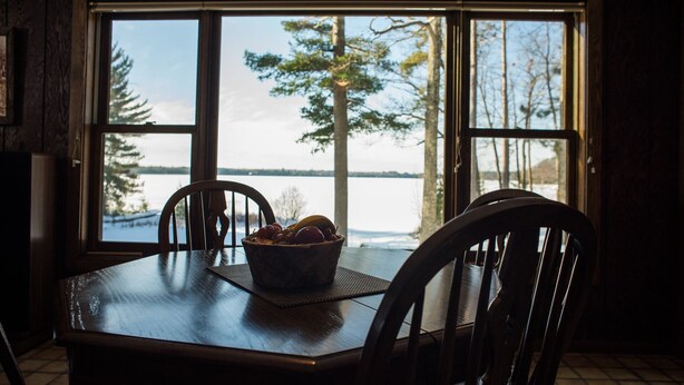 Una mesa de comedor con un frutero y dos sillas, ubicada frente a amplias ventanas con vistas al agua y árboles.