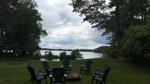 Un espacio al aire libre con asientos de césped, sillas tipo Adirondack y un brasero, con vista a un lago.
