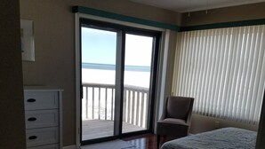 A bedroom with a bed, a chair, a dresser, and a view of the beach through a sliding door.