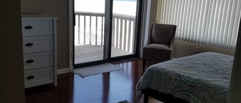Un dormitorio con cama, una silla, un aparador y vistas a la playa a través de una puerta corrediza.