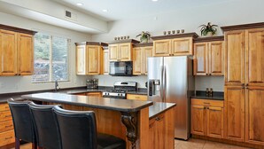 Una cocina con armarios de madera, un refrigerador de acero inoxidable, un microondas y una estufa. Hay dos taburetes negros y una ventana con persianas.