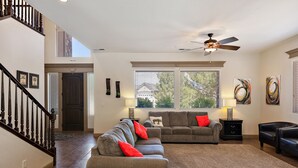 Amplia sala de estar con un sofá modular grande, un sofá más pequeño y un sillón de cuero negro. Se ve un ventilador de techo, y hay dos ventanas con persianas.