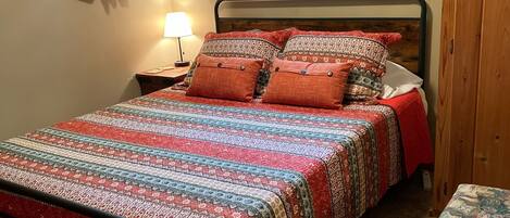 A bedroom with a bed, a nightstand, a lamp, a window with curtains, and a wooden door.