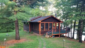 A cabin with a deck, surrounded by trees and a grassy area with stepping stones.