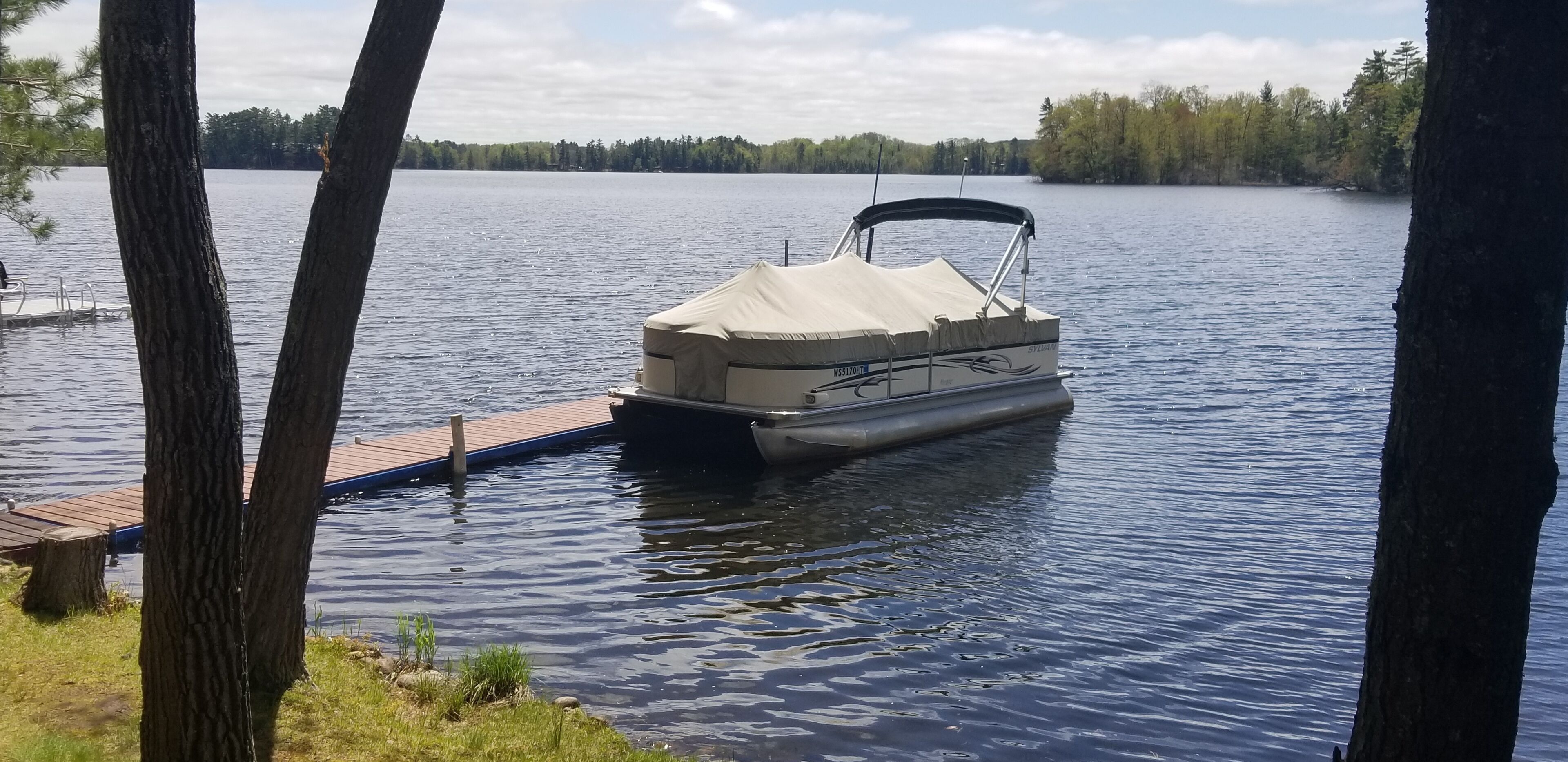 Een pontonboot aangemeerd aan een houten steiger, met een bebost landschap op de achtergrond.