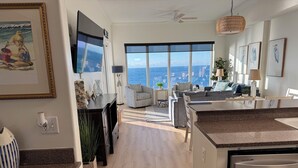 Ein modernes Wohnzimmer mit großem Fenster und Blick auf eine Stadtlandschaft, ein Küchenbereich mit Granit-Arbeitsplatten und ein Essbereich mit grauem Sofa.