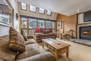 Un salon chaleureux avec une cheminée, un canapé agrémenté de coussins à carreaux, une table basse en bois et une vue sur un paysage enneigé par de grandes fenêtres.