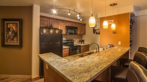 A kitchen with granite countertops, a stainless steel refrigerator, a microwave, and a sink. There are pendant lights above the counter and a framed picture on the wall.