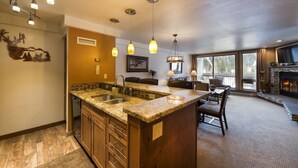 A spacious kitchen with granite countertops, a built-in sink, and a large island. There is a dining area with a fireplace and a large window offering a view of the outdoors.