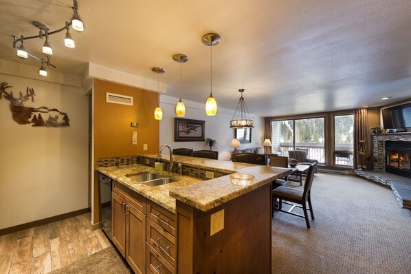 A spacious kitchen with granite countertops, a built-in sink, and a large island. There is a dining area with a fireplace and a large window offering a view of the outdoors.