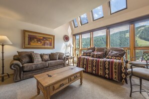 Un salon chaleureux avec un canapé en cuir marron, un canapé à motifs, une table basse en bois et une grande fenêtre donnant sur un paysage boisé.