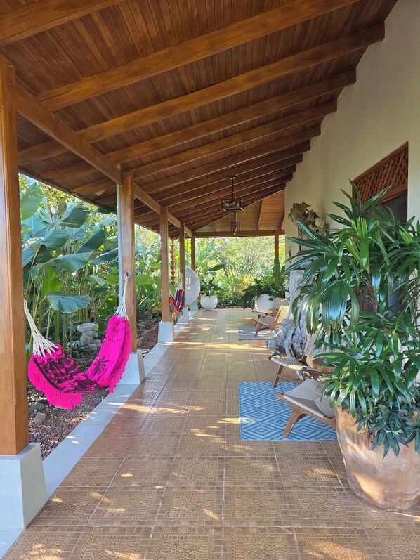 Un espacio al aire libre cubierto con suelo de baldosas, vigas de madera, sillones colgantes, plantas en macetas y una zona de estar.