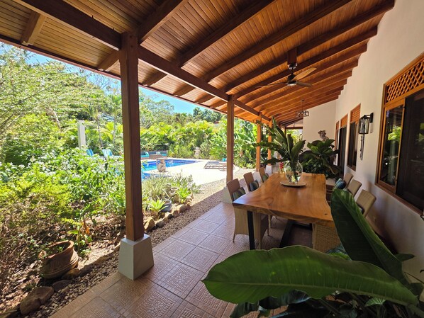 Un espace extérieur abrité, aménagé pour les repas, avec une piscine, un plafond en bois et des plantes tropicales.