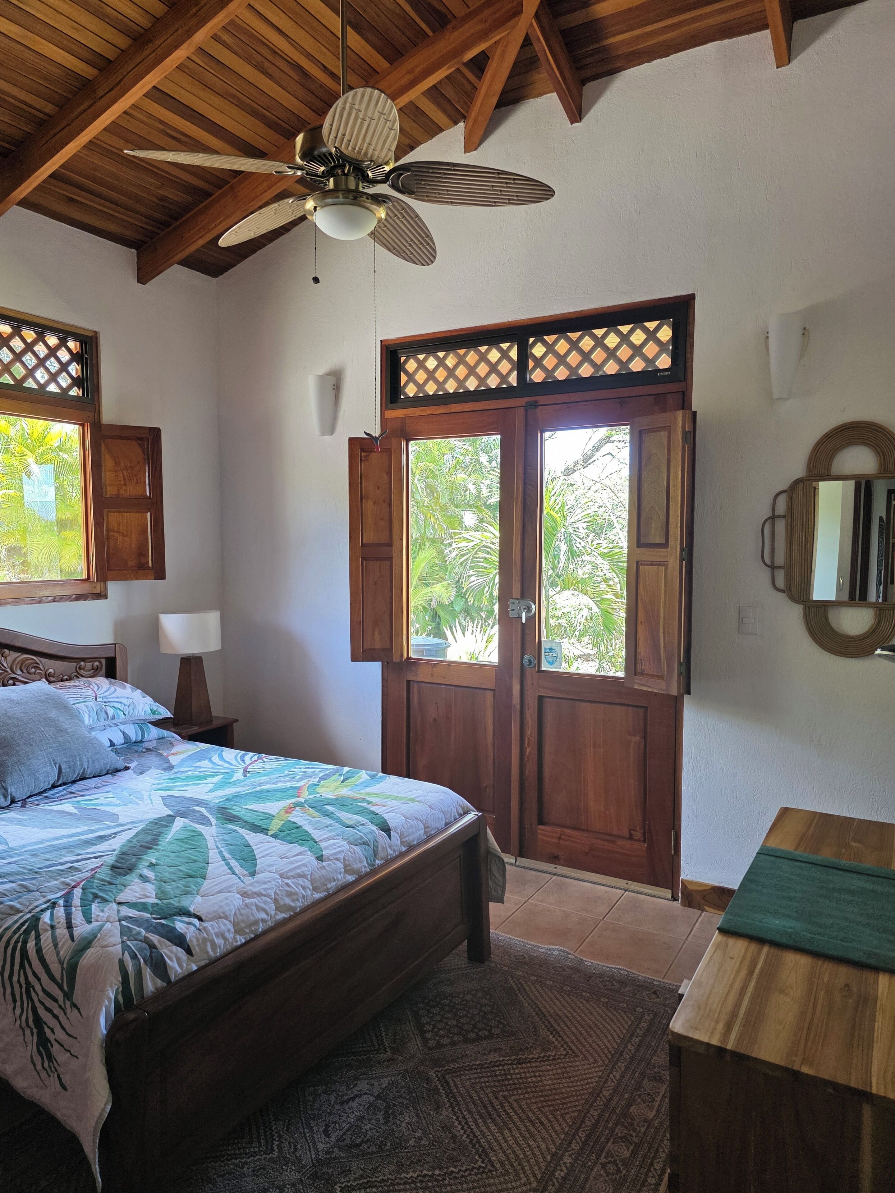 Une chambre à coucher avec un lit, un ventilateur de plafond en bois, une fenêtre donnant sur un paysage verdoyant et une porte en bois.