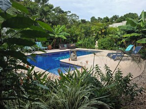 Una piscina con sillas de descanso y una escultura de piedra, rodeada de abundante vegetación.