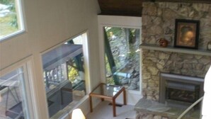 Un amplio salón con chimenea de piedra, un sofá y una mesa con lámparas. Grandes ventanales ofrecen vistas al exterior.