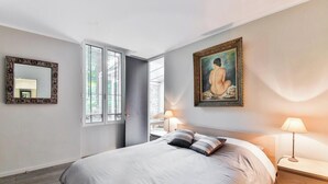 A bedroom with a bed, two bedside lamps, a painting on the wall, a mirror, and a window.