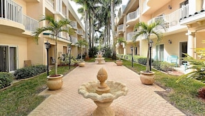 A hotel corridor with a central fountain, potted plants, and a tiled walkway.
