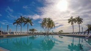 A pool with clear blue water, surrounded by palm trees and lounge chairs under a partly cloudy sky.