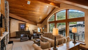 A spacious living room with a stone fireplace, leather furniture, and large windows offering a view of the surrounding landscape. - Plaza Lodge | 7 - 4 Bedroom Gold with private rooftop Jacuzzi (Vail)