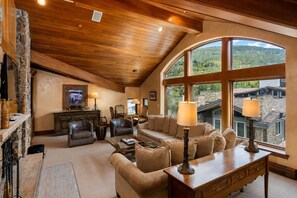A spacious living room with a stone fireplace, leather furniture, and large windows offering a view of the surrounding landscape. - Plaza Lodge | 7 - 4 Bedroom Gold with private rooftop Jacuzzi (Vail)