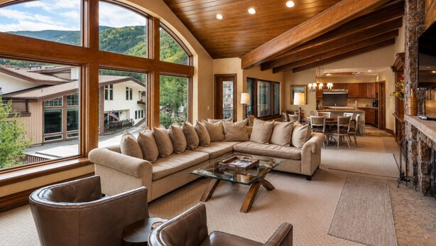 A spacious living area with a large window, a wooden ceiling, and a stone fireplace. - Plaza Lodge | 7 - 4 Bedroom Gold with private rooftop Jacuzzi (Vail)
