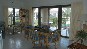 A spacious living area with a dining table and chairs, a bookshelf, and a sliding glass door leading to a balcony.