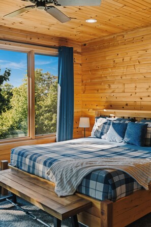 A wooden cabin bedroom with a large bed, a bench, a nightstand, a lamp, and a ceiling fan. - Northern Comfort Overlooking Caribou Lake- Spacious and Secluded (Lutsen)