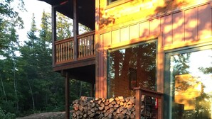 A wooden cabin with a deck, a stack of firewood, and a gravel path leading to the entrance.