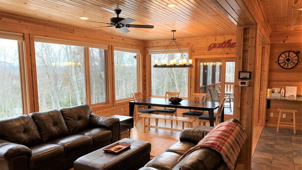 A cozy cabin interior with a leather sofa, dining table, and large windows offering a view of the forest.