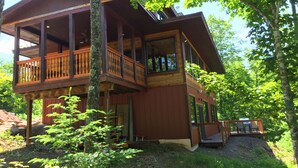 A two-story wooden cabin with a balcony, surrounded by trees and a gravel path.