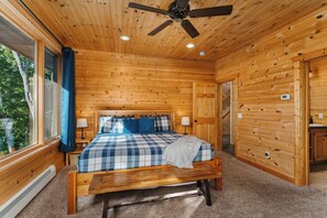 A wooden cabin bedroom with a bed, bedside lamps, a ceiling fan, and a view of the outdoors.