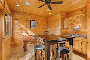 Interior de madera con una mesa de comedor, taburetes de bar, ventilador de techo y una zona de cocina con un letrero que ofrece bebidas gratis.