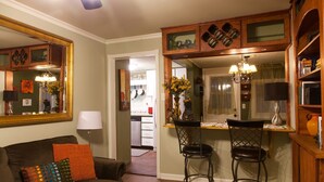 Una sala de estar acogedora con un sofá marrón oscuro, almohadas coloridas y una mesa de centro. Se ve una zona de cocina con armarios de madera y una mesa de comedor con dos sillas.