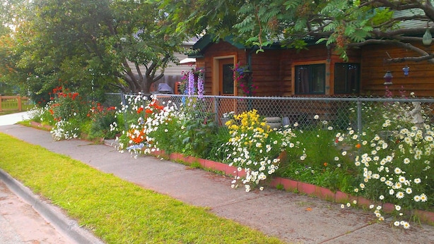 Una casa con fachada de madera, un porche delantero y diversos plantas con flores a lo largo de la acera.