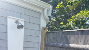 A white exterior wall with a metal outdoor shower, a wooden fence, and a brick pathway. - Great Location, Beach 4 minutes walk, Large Patio and Parking for one vehicle. (Dennis)