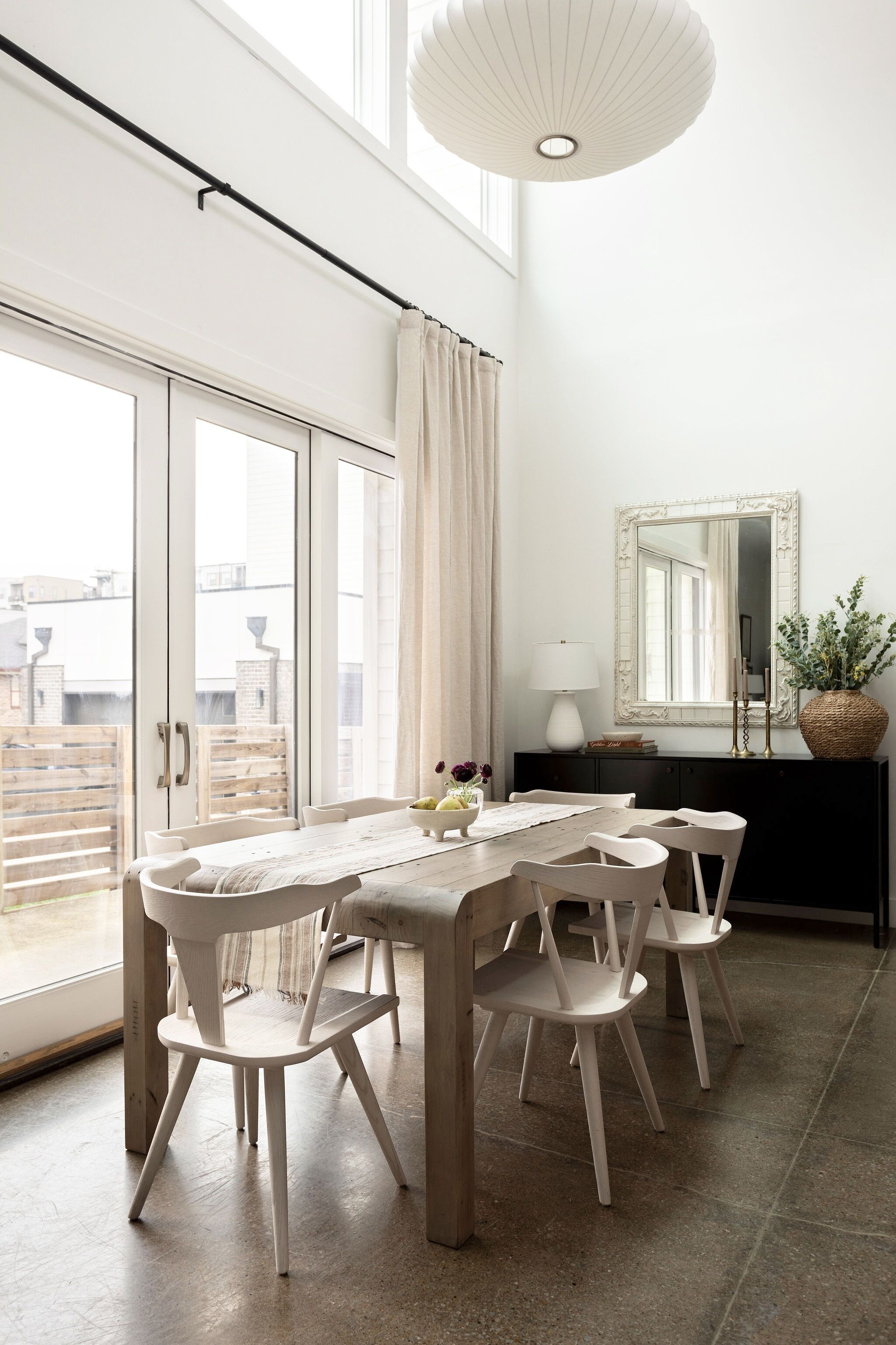 A dining area with a wooden table and white chairs, a large mirror, and a hanging light fixture.