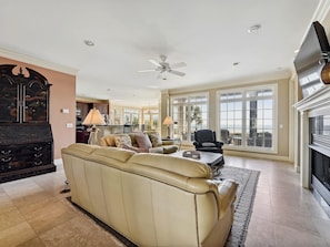 A spacious living room with a beige sofa, a fireplace, and large windows offering a view of the outdoors. - Luxury Oceanfront Home in North Forest Beach! (Hilton Head Island)
