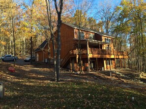 A two-story wooden cabin with a balcony, surrounded by trees and a gravel driveway.