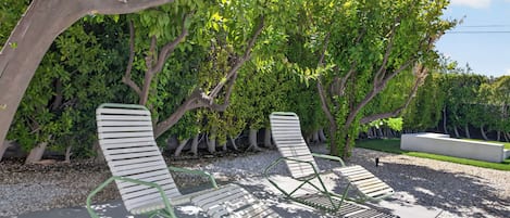 Twee buitenloungestoelen met metalen frames en witte kussens, geplaatst op een grindpad met een achtergrond van weelderige groene bomen.