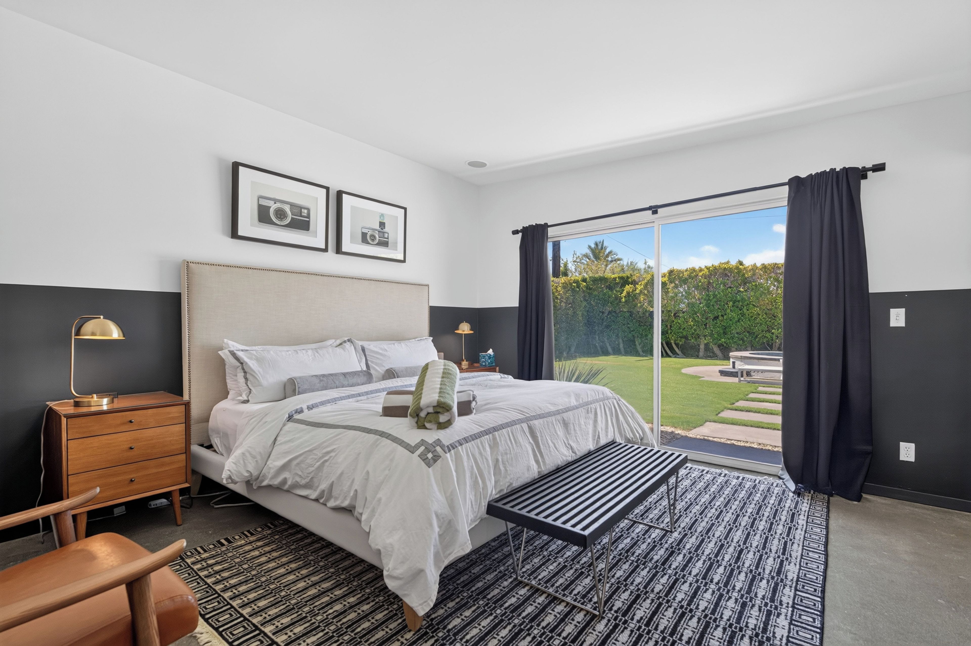 A bedroom with a bed, bedside tables, a desk, a chair, a window with curtains, and framed pictures on the wall.