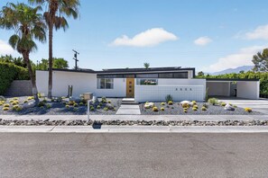 A modern two-story house with a flat roof, a yellow door, and a small garden with cacti and rocks.