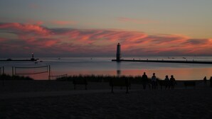 A beach scene with a lighthouse, calm water, and people sitting on benches. - Steps To The Beach & 2 Blocks To Downtown Frankfort - Sleeps 6-8 (Frankfort)