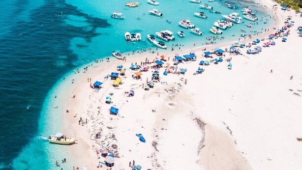 Une vue aérienne d’une plage où de nombreux bateaux sont amarrés, une plage de sable avec des parasols et des gens, et un ciel bleu dégagé.