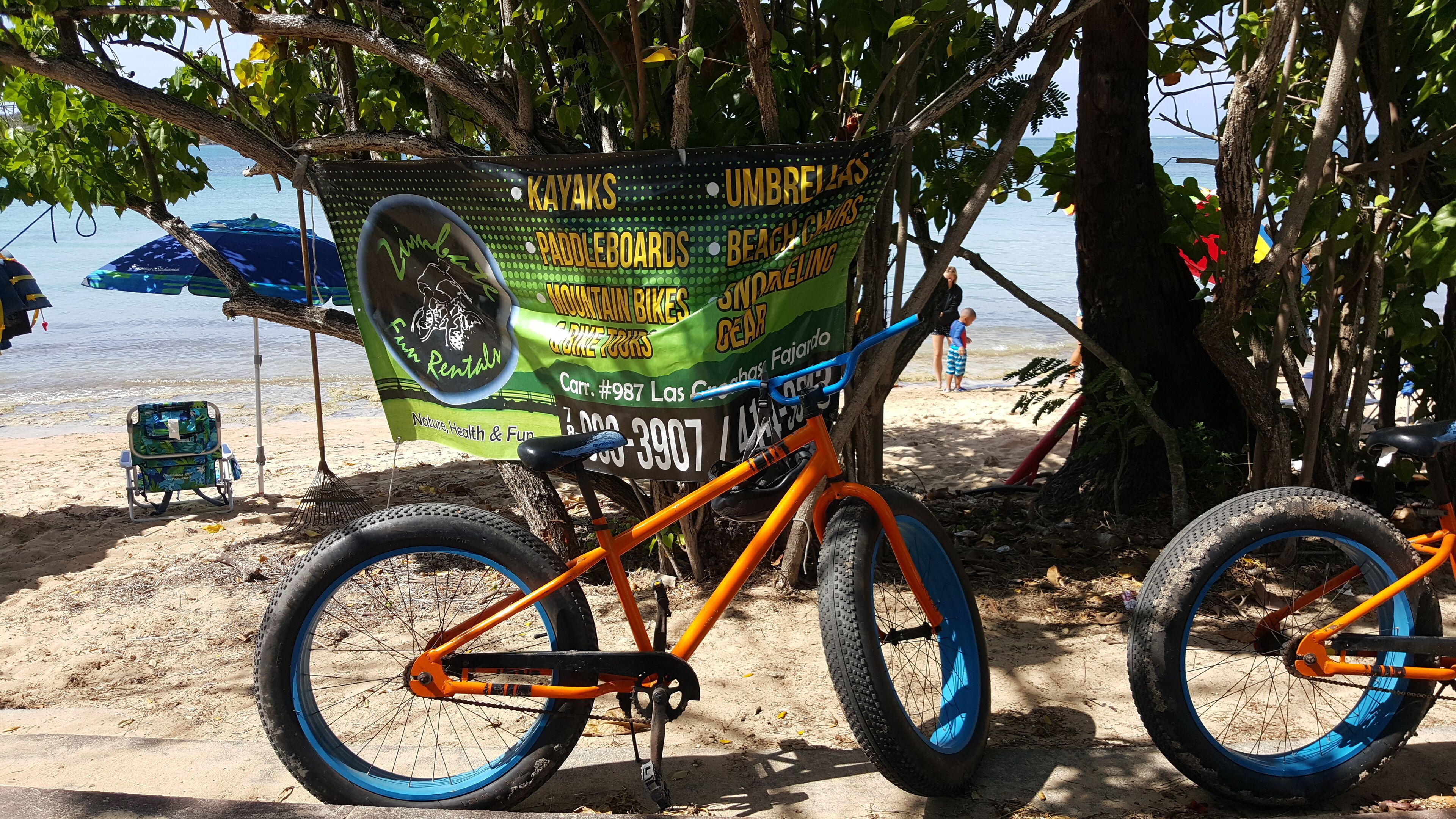 Escena de playa con una bicicleta y un cartel que anuncia servicios de alquiler como kayaks, sombrillas y sillas de playa.