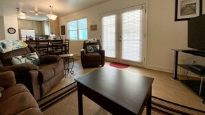 A living room with a sofa, armchair, coffee table, and a TV stand. There is a dining area with a table and chairs, and a kitchen in the background. - BEACHFRONT PRIVATE COTTAGE, FULL FLOOR, DECK, FREE PARKING BY ENTRANCE, 2BD 2BA  (Panama City Beach)